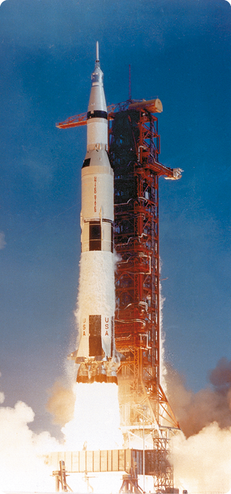 Fotografia de um foguete branco, com formato cilíndrico e a extremidade superior pontiaguda. Na extremidade inferior há chamas direcionadas para baixo e, ao redor fumaça. Ele está ao lado de uma estrutura vertical.
