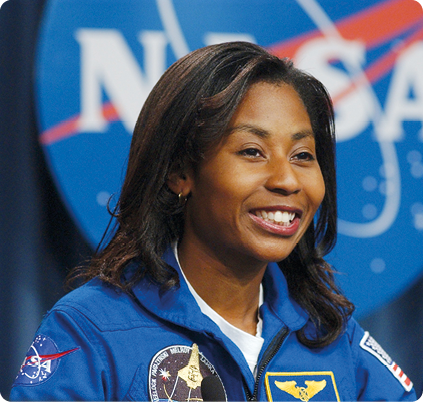 Fotografia do peito para cima de uma mulher negra sorrindo. Ela está usando roupa azul com logotipos, e ao fundo, uma placa azul com a seguinte sigla: 'NASA'.