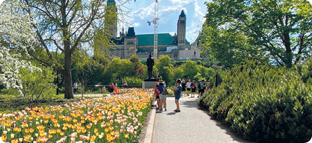 Fotografia de um local aberto, uma calçada ao centro com pessoas em pé e vegetação ao redor. À esquerda há um canteiro com flores coloridas. Ai fundo, uma grande construção com torres.