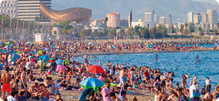 Fotografia de uma praia, com muitas pessoas usando roupa de banho na faixa de areia. À direita, o mar, com algumas pessoas dentro. Ao fundo, construções e prédios.