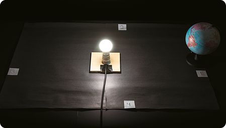 Fotografia do soquete com a lâmpada em cima das cartolinas pretas com os números de 1 a 4 e o globo terrestre inclinado ao lado. O ambiente está escuro e só há iluminação proveniente da lâmpada no dentro do modelo. 