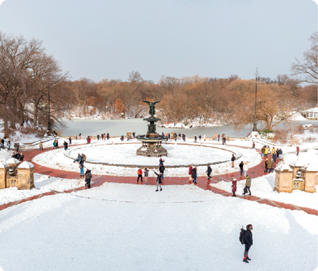Fotografia de um local aberto, ao centro uma fonte encoberta por neve, com uma estátua. Ao fundo um lago congelado e árvores sem folhas. Ao redor, neve no chão e pessoas. 