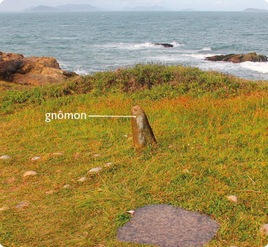 Fotografia de uma pedra comprida, em um local gramado, na posição vertical e com a ponta arredondada, próximo ao mar. Na pedra há a seguinte indicação: gnômon. Em volta dela há algumas pedras alinhadas no chão.