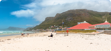 Fotografia de uma praia com larga faixa de areia e poucas pessoas nela. À esquerda, o mar, à direita construções, e ao fundo, montanhas.