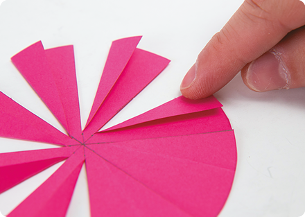 Fotografia. Destaque para uma mão dobrando as partes do círculo de cartolina cor-de-rosa, que foram previamente cortadas.