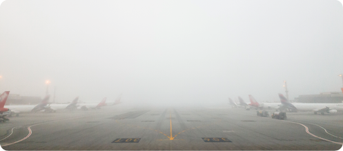 Fotografia. Vista de uma pista de um aeroporto com vários aviões estacionados. Há neblina entre as aeronaves.