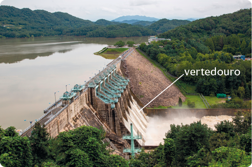 Fotografia. À esquerda, um lago artificial. À direita, uma parte da barragem com estruturas verticais e quedas de água. A água está caindo em um canal estreito com nível mais baixo. No local onde a água está escorrendo há a seguinte indicação: vertedouro.