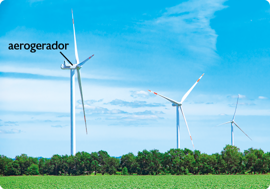 Fotografia. Vista de um campo gramado com árvores, e três aerogeradores, equipamento com uma haste vertical alta e três hélices em sua extremidade.