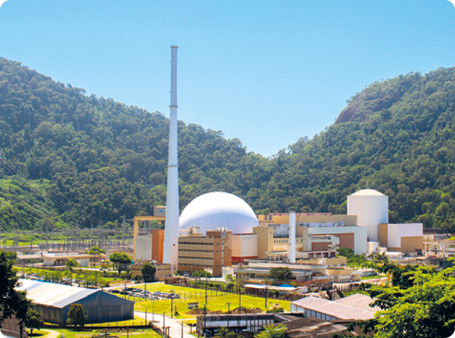 Fotografia. Vista de um local que apresenta vegetação, ruas e diversas construções, incluindo uma torre muito alta, uma construção em cúpula, prédios e estruturas cilíndricas, localizadas no centro.