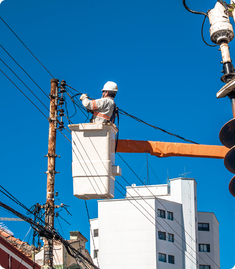 Fotografia. Um homem está dentro de uma plataforma elevada que está presa a uma haste horizontal. Ele está usando luvas, capacete e um macacão, e está manuseando fios elétricos que estão no alto de um poste.