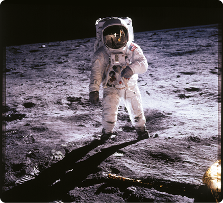 Fotografia de um astronauta em pé, sobre uma superfície rochosa. Ele usa um traje branco grande, botas, luvas, um objeto retangular similar a uma mochila em suas costas e um capacete fechado cobrindo toda a cabeça, com uma viseira reflexiva.
