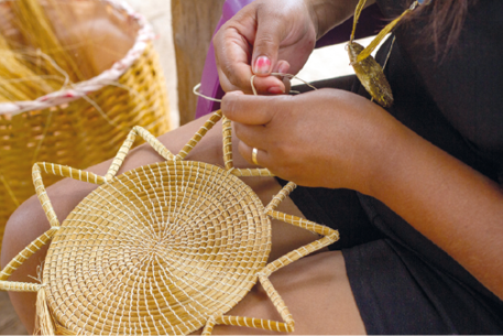 Fotografia. Duas mãos de uma pessoa manuseando um fio dourado de capim. Sobre as pernas dessa pessoa, uma peça dourada composta por círculo com triângulos ao redor. Ao fundo, há um cesto com mais fios dourados de capim.