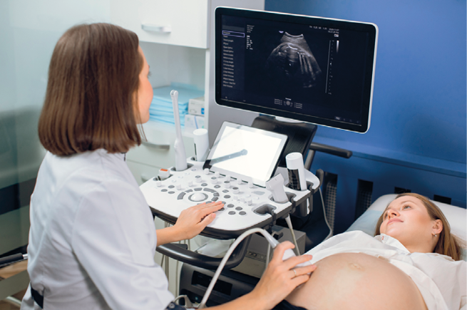 Fotografia. À esquerda, uma mulher está sentada olhando para um visor, com a mão esquerda sobre os botões de um aparelho que está à sua frente. Com a mão direita, ela segura um instrumento sobre a barriga de outra mulher, que está deitada à sua direita. No visor, é projetada uma imagem em branco e preto do interior da barriga da mulher deitada.