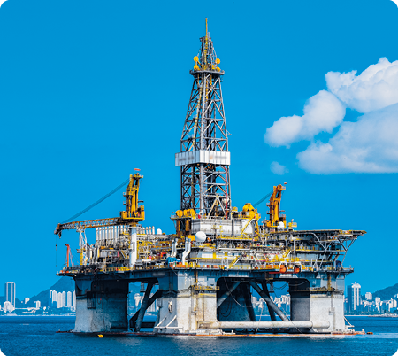 Fotografia. Uma plataforma de petróleo. Ela está em meio ao oceano e possui estruturas de concreto com uma torre metálica alta, na posição vertical, além de guindastes e outras estruturas metálicas ao lado. Ao fundo, há prédios e montanhas.