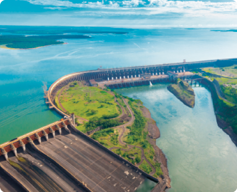 Fotografia. Vista aérea de um local com uma região com água em nível alto e outra região com água em nível mais baixo, separadas por uma barragem composta por um muro com estruturas verticais. Em um local da parede há uma estrutura com formato de rampa, com a parte baixa na direção do trecho de água no nível mais baixo. Há trechos de vegetação na parte frontal e na parte posterior.