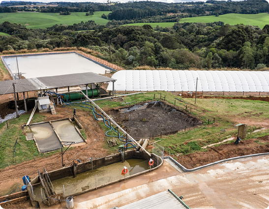 Fotografia. Vista aérea de uma área gramada com tanques contendo líquido e um buraco quadrado contendo um material preto. Atrás, há uma construção com telhado e um cilindro abaixo, e outra construção com uma cobertura branca. No fundo, há outro tanque maior contendo líquido e muitas árvores ao redor.