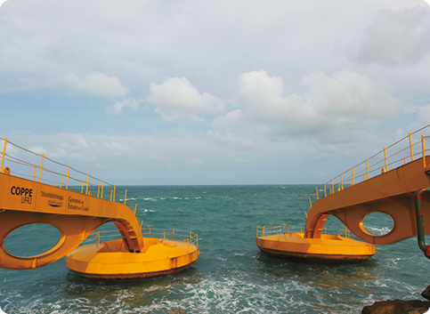 Fotografia. Duas hastes metálicas horizontais grandes, ligadas a bases circulares que estão sobre a superfície do mar.