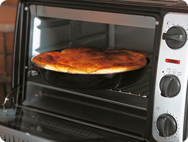 Fotografia. Uma forma escura dentro de um forno elétrico com grades no interior e botões na lateral. A forma contém um alimento que está assado.