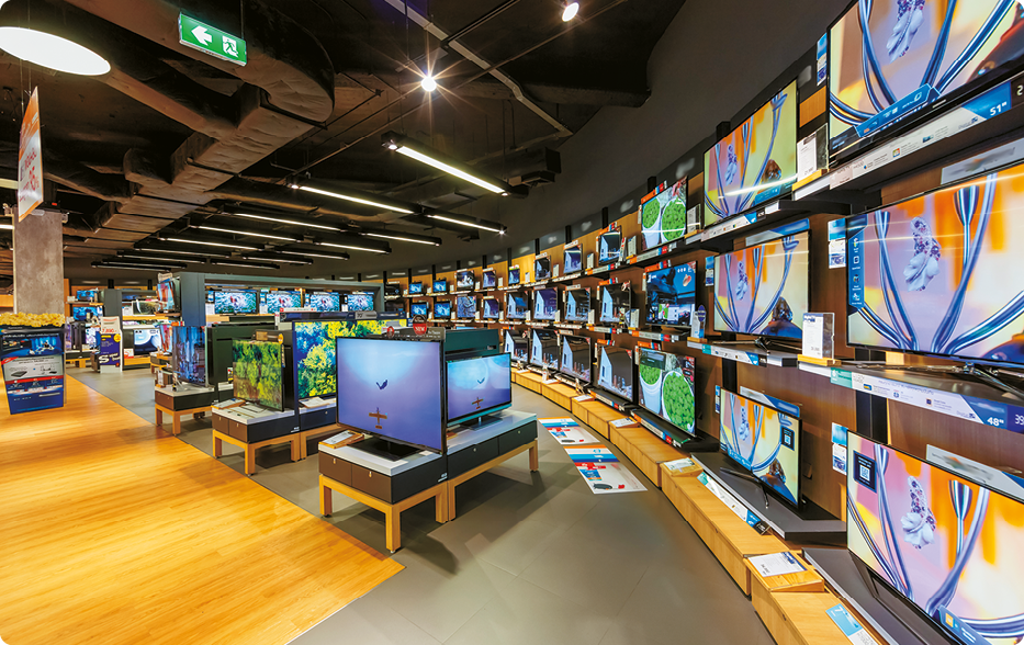 Fotografia. Um local fechado com muitos televisores em prateleiras próximas a uma parede. No centro do local, há mesinhas com televisores em cima. Todos os televisores estão ligados.