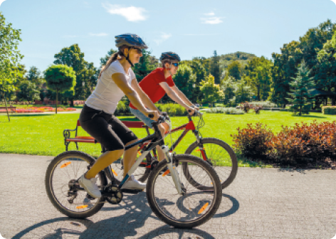 Fotografia. Duas pessoas andando de bicicleta, lado a lado. Elas estão usando capacete e óculos escuros em um local que tem uma pista e gramado com plantas. Ao fundo, há muitas árvores.