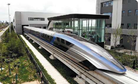 Fotografia. Um trem pontiagudo, que corre sobre uma superfície elevada que sai de uma passagem em um edifício. Abaixo, há trechos de grama, e ao lado, outro prédio.