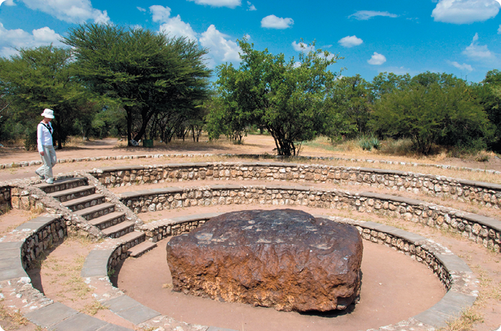 Fotografia. Em um local aberto, com árvores e vegetação rasteira, há um meteorito retangular de tonalidade amarronzada no chão. Ele está posicionado no centro, rodeado por areia e cercado por muros circulares baixos de pedra. À esquerda, há uma pessoa descendo uma escada em direção ao meteorito.