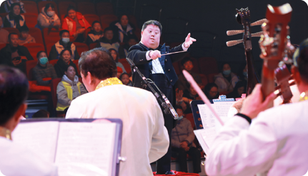 Fotografia de um maestro fazendo gestos para os músicos de uma orquestra. Ele tem a cabeça grande, rosto arredondado, nariz pequeno e olhos oblíquos.