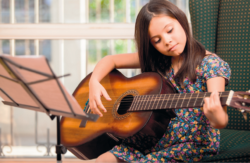 Fotografia de uma criança sentada, segurando um violão, a mão direita está sobre as cordas e a mão esquerda segurando o braço do violão; ela está olhando para o violão e a sua frente há um suporte com um papel.