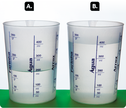 Fotografia. Dois recipientes estão sobre uma superfície verde. Ambos possuem capacidades que variam de 50 a 500 mililitros. Nas laterais dos recipientes está escrito água. Marcado com a letra A, um copo está preenchido com água até a marca de 200 mililitros. Marcado com a letra B, o outro copo está preenchido com água até a marca de 400 mililitros.