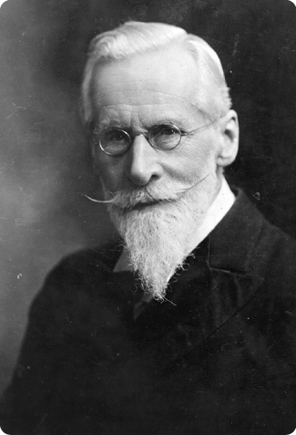 Fotografia em preto e branco do químico William Crookes, do peito para cima. Ele tem cabelo curto, uma barba e um bigode grandes. Está vestindo uma camisa clara, um casaco escuro e óculos redondos.