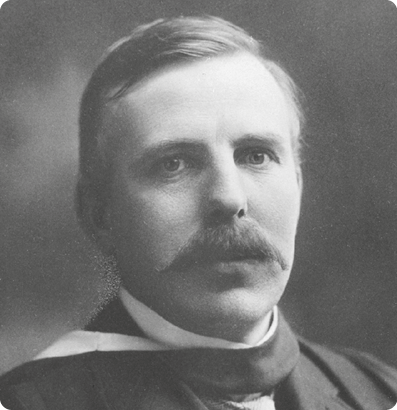 Fotografia em preto e branco do cientista Ernest Rutherford, dos ombros para cima. Ele tem cabelo curto e um bigode. Está vestindo um casaco escuro.