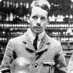 Fotografia em preto e branco do físico Henry Gwyn Jeffreys Moseley, da barriga para cima. Ele possui cabelo curto, bigode e está vestindo uma camisa clara, uma gravata escura e um casaco.