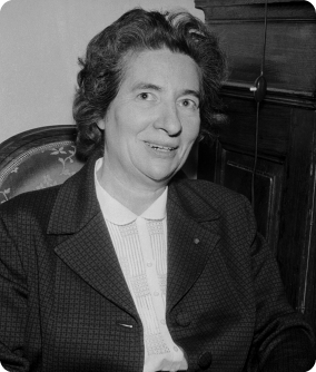 Fotografia em preto e branco da química Marguerite Perey, da barriga para cima. Ela possui cabelo curto e está vestindo uma camisa clara e um paletó escuro.