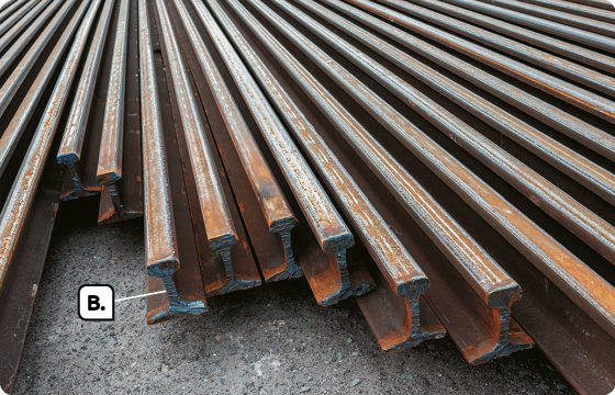 Fotografia. Várias barras de metal com manchas alaranjadas e marrons. Indicada com a letra B, a ponta de uma das barras com coloração cinza.