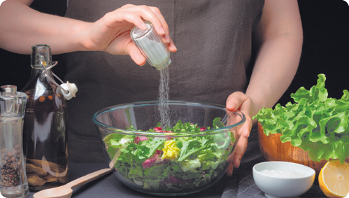 Fotografia de uma pessoa do peito para baixo adicionando sal a uma salada com folhas verdes, amarelas e vermelhas, utilizando um saleiro. A salada está dentro de um recipiente arredondado transparente. À esquerda, há um frasco com azeite e outro com pimenta. À direita, há outro recipiente com folhas verdes, um com sal e um limão cortado ao meio.