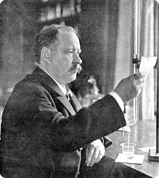 Fotografia em preto e branco do químico Svante August Arrhenius, da cintura para cima, olhando para um pedaço de papel em seu laboratório. Ele tem cabelo curto, um bigode e está vestindo uma camisa clara e um paletó escuro. Ao seu lado, há um objeto cilíndrico de vidro alongado preso a uma haste com base quadrada, e abaixo dele, um recipiente cilíndrico transparente com uma boca achatada, ambos posicionados sobre uma mesa.