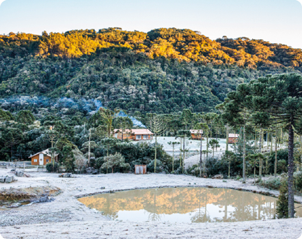 Fotografia. Vista de um local com um lago no centro, rodeado por gelo. Ao fundo, há árvores e algumas casas cobertas de gelo. Mais distante, há uma montanha com vegetação.