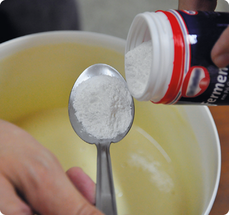 Fotografia. Duas mãos de uma pessoa, uma despejando o pó branco de um recipiente de fermento em uma colher segurada pela outra mão. A colher está sobre um recipiente redondo com massa viscosa amarela.