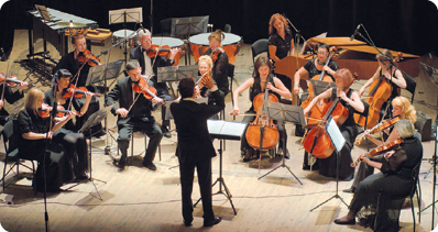 Fotografia de pessoas sentadas em cadeiras tocando instrumentos musicais em um palco, em volta de uma pessoa em pé com os braços levantados.