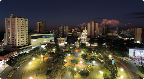 Fotografia. Vista aérea de uma praça com árvores e ruas com carros, durante a noite. Vários postes de iluminação estão espalhados pelo local, mantendo o local bem iluminado. Nas laterais da praça, há prédios e outras construções.