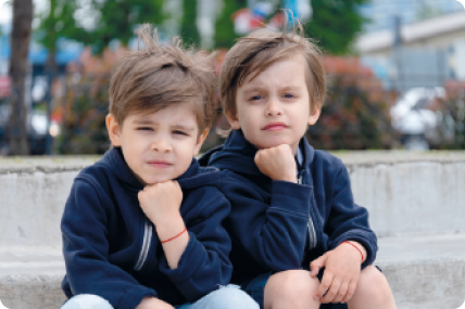 Fotografia de dois meninos de cabelos curtos, sentados lado a lado, com a mão fechada sob o queixo, olhando para frente, usando blusas de frio iguais. Eles são parecidos, mas um deles tem um rosto mais arredondado que o outro e as sobrancelhas mais compridas.