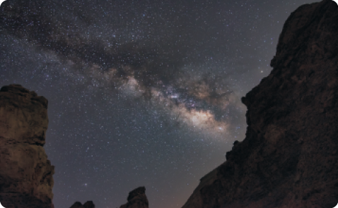 Fotografia de céu escuro estrelado, com uma região dele formada por uma faixa brilhante e difusa, com algumas porções mais escuras. Ao redor, partes de montanhas.
