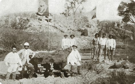Fotografia em preto e branco de pessoas ao lado de um meteorito, uma grande rocha. As pessoas estão vestindo calça, camisa comprida, botas e chapéus. Ao redor há vegetação rasteira e árvores.