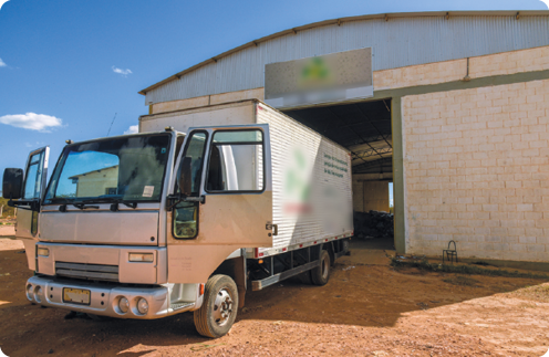 Fotografia de um caminhão baú estacionado de ré, em frente à entrada de um galpão, com as portas abertas.