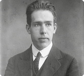 Fotografia em preto e branco do físico Niels Bohr, do peito para cima. Ele tem cabelo curto e está vestindo uma camisa clara, uma gravata escura com bolinhas claras e um paletó escuro.