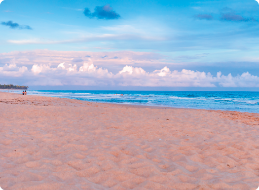 Fotografia de uma praia. Local com muita areia e o mar ao fundo. Acima, há várias nuvens e o céu azul.