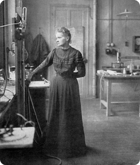 Fotografia em preto e branco da física Marie Curie, em pé em seu laboratório. Ela possui cabelo curto e está vestindo um vestido longo escuro. Ela está em frente a um objeto alto e retangular com hastes. Ao fundo, há uma mesa com objetos em cima.