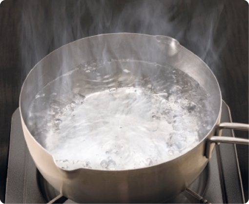 Fotografia. Uma panela sobre um fogão com água fervendo. Há bolhas na superfície da água e acima dela há uma névoa branca.