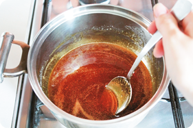 Fotografia. A mão de uma pessoa segurando uma colher e mexendo um líquido amarronzado dentro de uma panela redonda sobre o fogão.