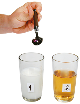 Fotografia. Dois copos lado a lado. No copo marcado com o número 1, há um líquido branco. No copo marcado com o número 2, há um líquido amarelado. Em cima dos copos há uma mão segurando uma colher com um líquido roxo.
